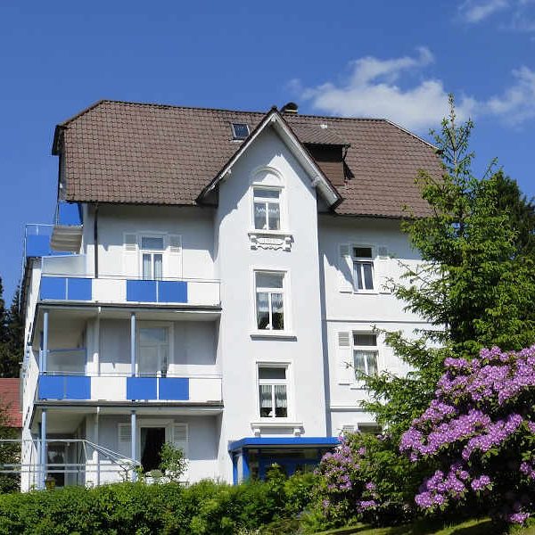 Frontansicht Hotel Fidelitas Weißes Haus mit blauen Balkonen, umgeben von Bäumen und blühenden Sträuchern.